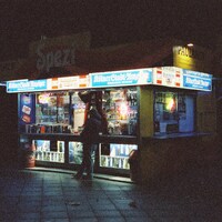 A person stands at a brightly lit kiosk at night by Maximilian Bungart