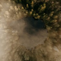 Aerial view of a dark lake surrounded by foggy forest by Aivars Vilks