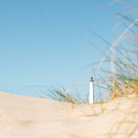 A white lighthouse behind sand dunes with tall grass under a clear sky by Max Böhme