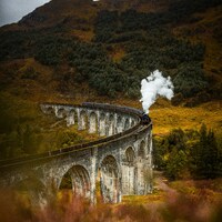 Steam train crosses a stone viaduct in a scenic landscape by David Becker
