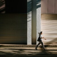 Woman walking on sidewalk with building shadows by Takashi S