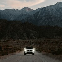 White truck drives on a dirt road towards mountains by Royce Fonseca