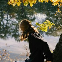 A woman in a black sweatshirt by water under yellow oak leaves by Alina Chernovolova