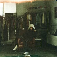Person in fluffy hat sits in armchair surrounded by clothes by Zach Miller