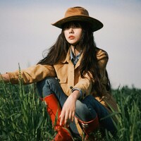 Woman in hat and boots squats in a green field by 光术 山影