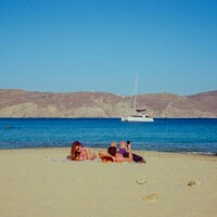Two people sunbathing on a sandy beach with a sailboat by Jean Carlo Emer